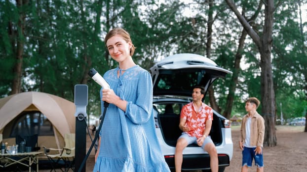 Outdoor adventure and family vacation camping at sea travel by eco friendly car. Cheerful woman or mother holding, pointing EV charger point with playful and happiness posture in campsite. Perpetual