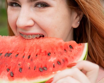 Close-up portrait of red-haired young woman with braces eating watermelon outdoors.