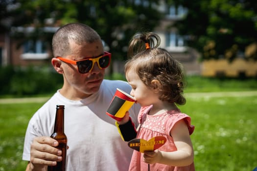 Portrait of a handsome young caucasian male father in sunglasses and baby daughter drinking drinks and celebrating belgium day at a picnic in a city park on a sunny summer day, close up side view.