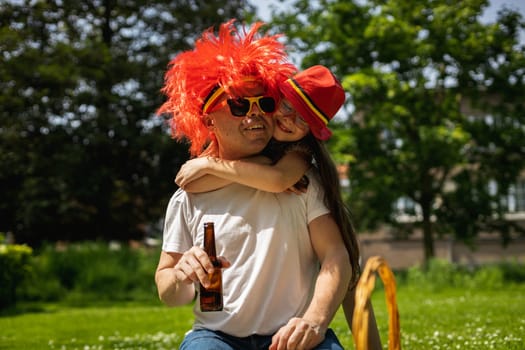 Portrait of handsome young caucasian man in sunglasses with belgian flag, red wig holding bottle of beer in hands and happy daughter hugging his neck, sitting on lawn and celebrating belgium day in city park on sunny summer day, close up side view.