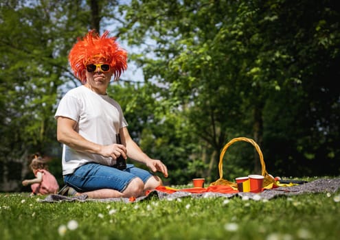 Portrait of a handsome young caucasian man in Belgian flag sunglasses, red wig with a beer from a glass bottle in his hands, sitting on the lawn and celebrating belgium day in a city park on a sunny summer day, close-up side view.