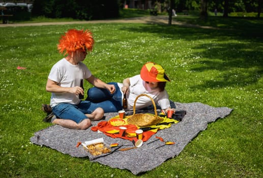 Portrait of a handsome young caucasian couple in Belgian flag sunglasses, red wig and hats holding a bottle of beer in their hands and sitting on the lawn celebrating belgium day in a city park on a sunny summer day, top side closeup view.