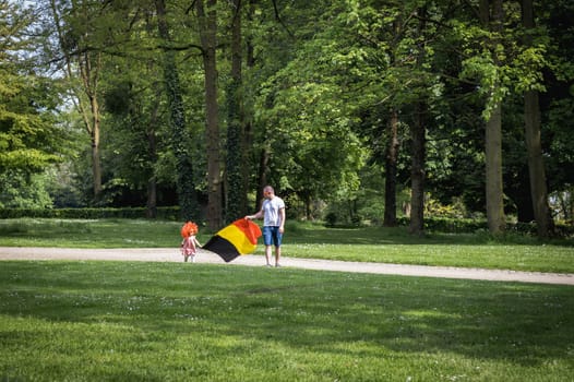 Portrait of one beautiful Caucasian young father with his little daughter in a pink dress with a red wig, their heads carry the Belgian flag together while walking along the lawn in the park on a summer day, side view close-up.
