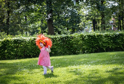 Portrait of one beautiful Caucasian little girl in a pink dress with a red Belgian flag wig on her head runs across the lawn in the park on a summer day, side view close-up with copy space.