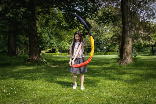 Portrait of one beautiful Caucasian little brunette girl in a skirt with her hair down, twirling a twisted Belgian flag in a circle in a city park on a summer day, close-up side view.