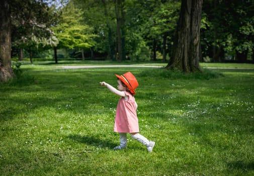 A portrait of one beautiful Caucasian little baby girl wearing a red Belgian flag hat stands sideways on the lawn in a city park on a summer day and points her finger to the left, close-up side view.