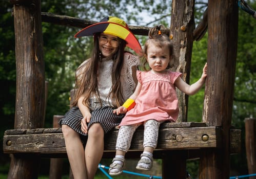 Portrait of two beautiful Caucasian happy little brunette girls in a Belgian flag hat sitting on a wooden walkway of a rope swing in a city park on a summer day, close-up side view.