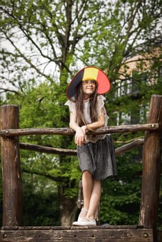 Portrait of one beautiful Caucasian brunette girl with glasses and a Belgian flag hat, looking to the side with a smile and standing at full length on a wooden bridge on a playground in a city park, close-up view from below.