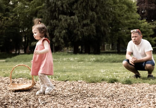 Portrait of one beautiful Caucasian little girl in a pink dress and with a ponytail on the top of her head standing near a wicker basket looking to the side, and a father is squatting next to him and looking after his daughter in the park on a summer day, close-up side view.