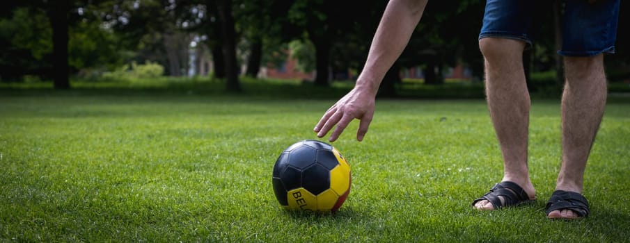 An unrecognizable young man Caucasian man reaches for a soccer ball with Belgium written on it, lying on the green lawn of a city park on a summer day, close-up view from the side.