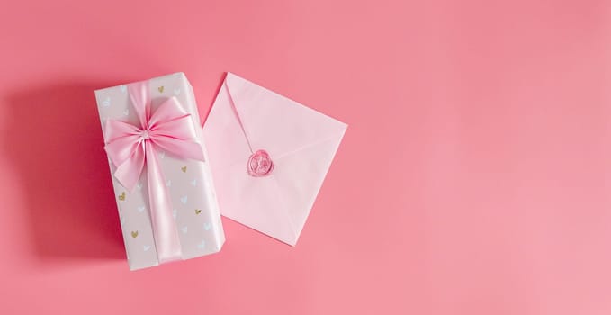 One beautiful gift box with a bow and a sealed envelope lie on the left on a pink background with copy space on the right, flat lay close-up.