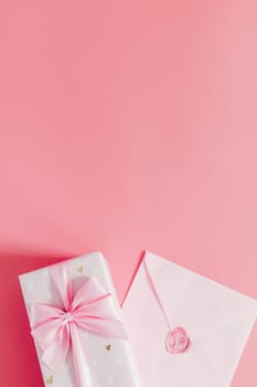 One beautiful gift box with a bow and a sealed envelope lie below on a pink background with copy space on top, flat lay close-up.