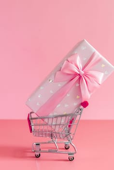 One large beautiful gift box with a bow lies in a mini shopping cart on the right on a pink background, close-up side view.