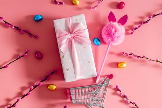 One large beautiful gift box with a bow, a fluffy bunny handle, chocolate Easter eggs in multi-colored wrappers and willow branches fall out of a mini shopping cart on a pink background, flat lay close-up.