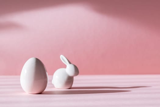 Porcelain figurines of Easter eggs and bunny stand on the left on a pink background with shadows and copy space on the right, side view close-up.