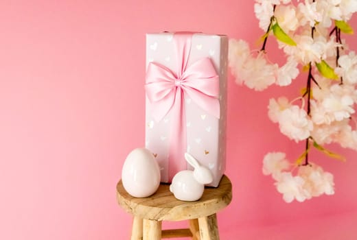 Porcelain Easter egg, bunny, large gift box with a bow stand on a small homemade decorative wooden stool on a pink background with a blurred branch of an apple tree blossom, side view close-up with selective focus.