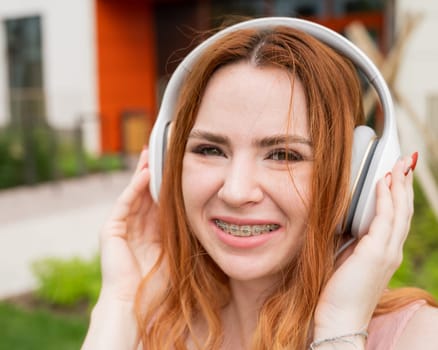 Portrait of a young red-haired woman with braces on her teeth listening to music on headphones outdoors