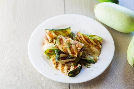 Grilled zucchini pieces with garlic sauce in a white plate. Light gray wooden background.