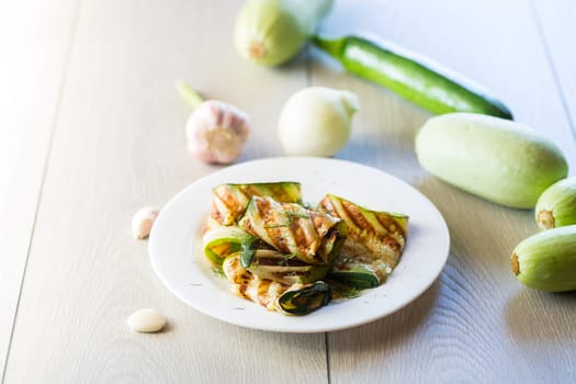 Grilled zucchini pieces with garlic sauce in a white plate. Light gray wooden background.