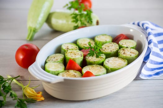 Raw zucchini with tomatoes prepared for baking in a ceramic form, on a wooden table.