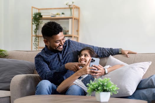 a couple sitting on the sofa looking at their mobile phones smiling and laughing together during happy holiday time.