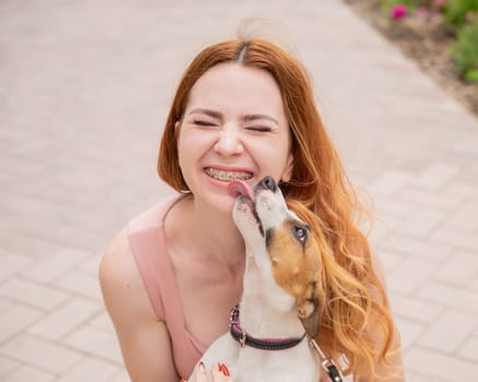 Dog jack russell terrier licks the owner in the face outdoors. Girl with braces on her teeth