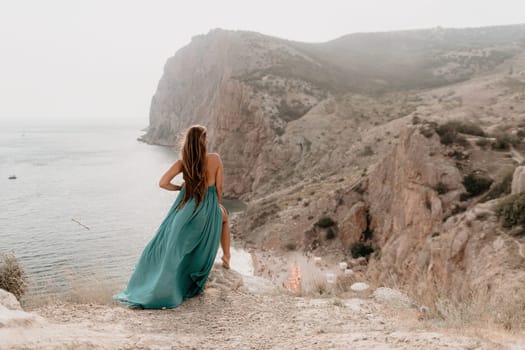 Side view a Young beautiful sensual woman in a mint long dress posing on a volcanic rock high above the sea during sunset. Girl on the nature on overcast sky background. Fashion photo