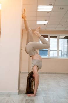 The girl is training in the yoga studio. Dressed in a beige tracksuit doing a handstand against the wall. Healthy lifestyle and yoga concept