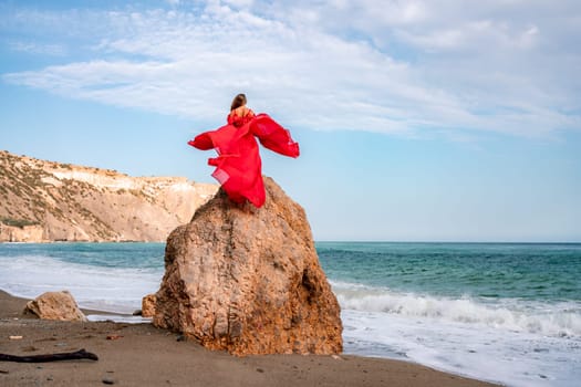 woman sea red dress. Woman with long hair on a sunny seashore in a red flowing dress, back view, silk fabric waving in the wind. Against the backdrop of the blue sky and mountains on the seashore