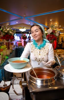 A joyful Uzbek woman in ethnic attire and ornate accessories happily serves soup from a sizable metal tureen at a lavish banquet adorned with vibrant floral decorations..