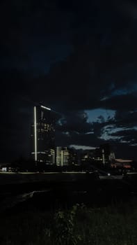 A tall skyscraper stands out among other buildings in the city at night. The sky is dark and cloudy, with a hint of light on the horizon.