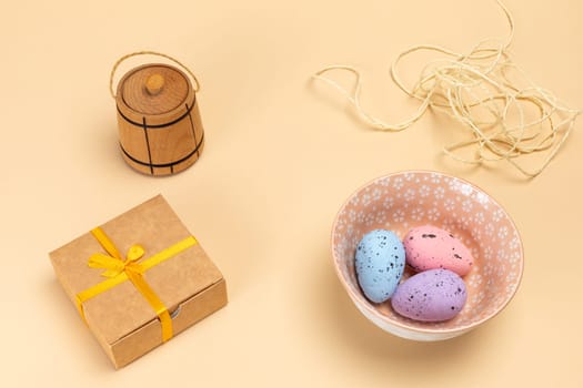 Bowl with colored Easter eggs, a small wooden barrel, a gift box and a rope on the beige background. Top view.