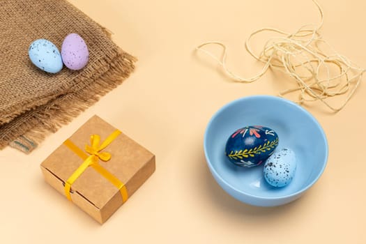 Blue bowl with colored Easter eggs, a sackcloth bag, a gift box and a rope on the beige background. Top view.