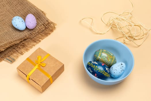 Blue bowl with colored Easter eggs, a sackcloth bag with eggs, a gift box and a rope on the beige background. Top view.