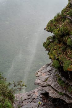 High waterfall on cliff with wind-spread mist over lush forest.