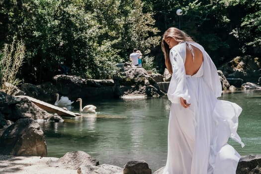 a beautiful woman in a long white dress looks into the distance at a beautiful lake with swans