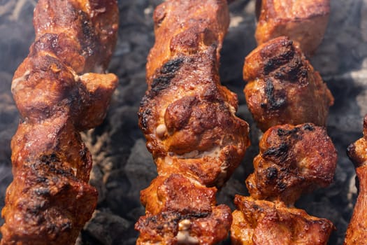 Tasty juicy pork barbecue cooking on metal skewers on charcoal outdoors grill with fragrant fire smoke. Cooking during summer picnic. Close-up view, selective focus on pieces of delicious roast meat.