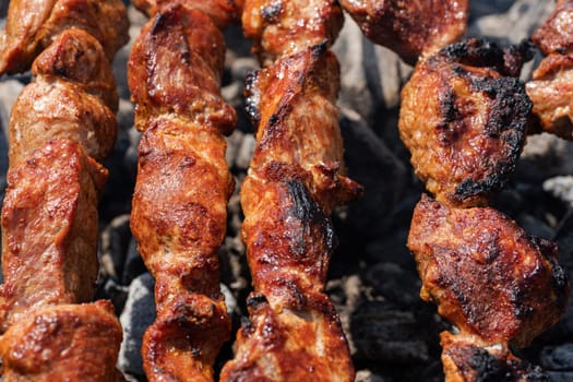 Cooking during summer picnic - appetizing juicy pork shish kebabs on outdoors metal skewers on charcoal grill with fragrant fire smoke. Close-up view, selective focus on tasty pieces of roast meat
