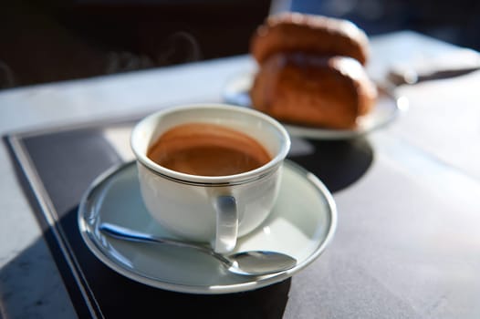 Still life. Elegant white ceramic cup of freshly brewed espresso coffee with foam on a sauce, blurred crispy croissants on the background, on the table of a sidewalk cafe. French bakery and patisserie