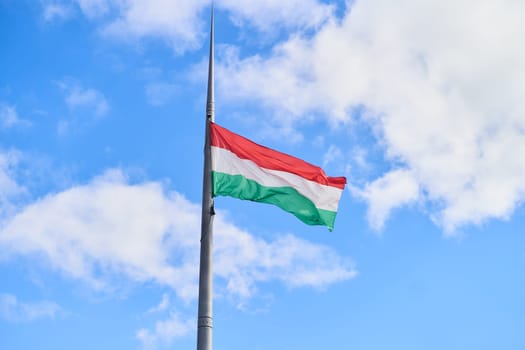 A Hungarian flag flutters in the wind against a blue sky with cumulus clouds. The flag is attached to a pole near a tree