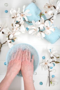 woman soaks her hands in a bowl with soapy water and sea pebbles for a delicate manicure spa procedure in the salon decorated with magnolia flowers, High quality photo