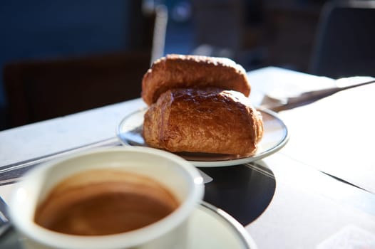 Two freshly baked French crispy croissants with chocolate cream on a plate and cup of coffee on the blurred foreground, served for breakfast in French traditions. Cuisine. Culinary. Bakery. Patisserie