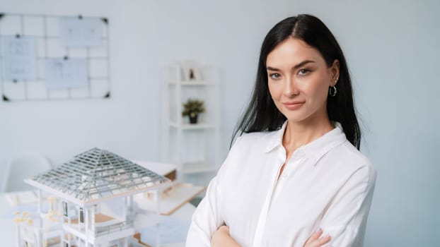 Close up portrait of young beautiful professional architect engineer cross arms and poses confidently with house model at white modern office with blueprint. Creative living and design. Immaculate.