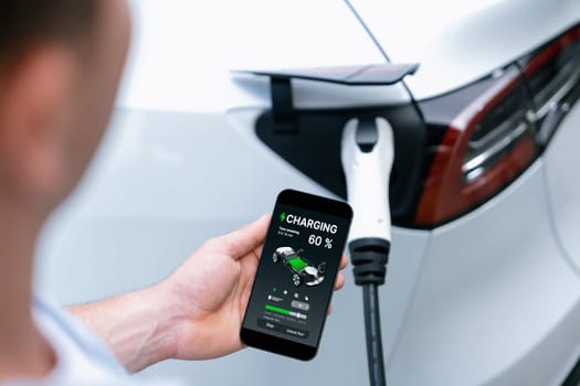 Modern eco-friendly man recharging electric vehicle from EV charging station, using Innovative EV technology utilization for tracking energy usage to optimize battery charging on smartphone.Synchronos