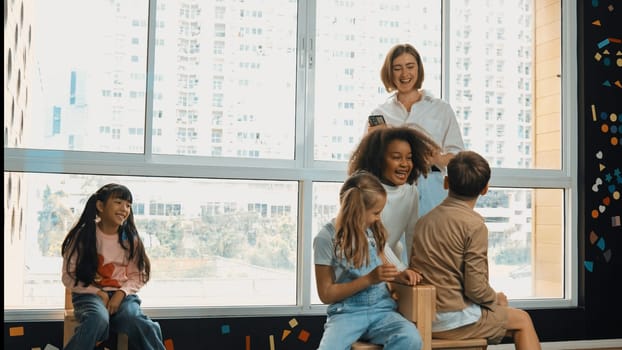 Diverse children playing musical chairs games while young smart beautiful teacher turn off music. Multicultural students running and sitting on chair in creative activity. Music lesson. Erudition.