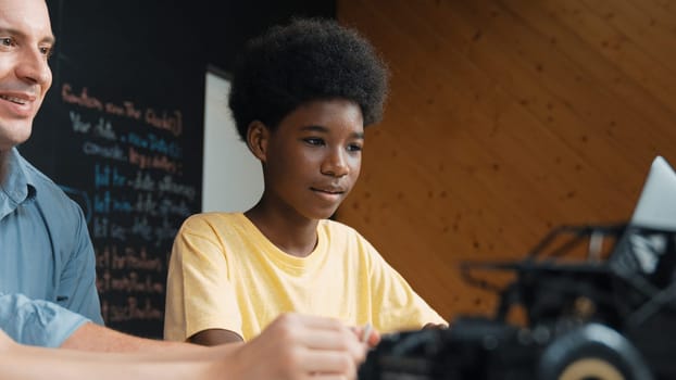 African boy using laptop to program code or prompt with car model placed while teacher and caucasian friend study about robotic construction. Student study about car model structure. Edification.