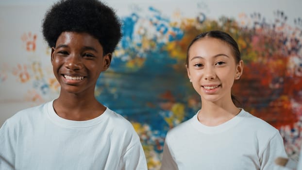 Smart african boy and caucasian girl look at camera while standing with confident at colorful stained wall. Teenager with mixed races pose at camera while hold paintbrush and palette. Edification.