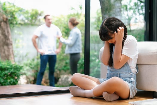 Stressed and unhappy young girl huddle in corner, cover her ears blocking sound of her parent arguing in background. Domestic violence at home and traumatic childhood develop to depression. Synchronos