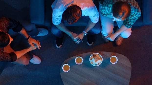 Happy family and colleague sitting at dark living room with red filter while cheering football team together. Caucasian people enjoy watching tv and giving high five and clinking glasses. Convocation.