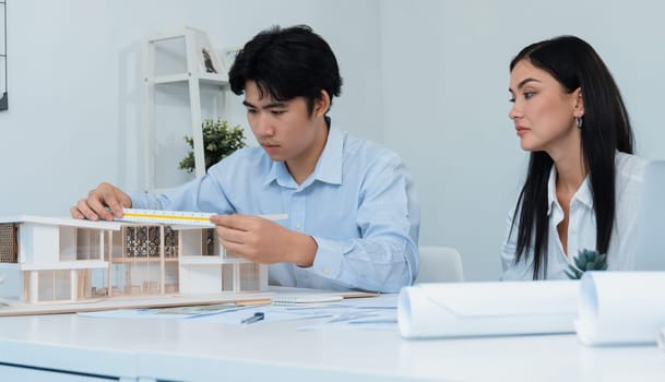 Professional male asian architect using ruler to measure house model length while young beautiful caucasian colleague using laptop to analyzed data on meeting table with house model. Immaculate.